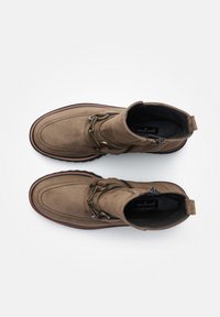 Bruine suède enkelboots met een decoratieve metalen kettingdetail, ronde neus, zijrits en contrasterende zool. De binnenkant heeft een zwarte voering.