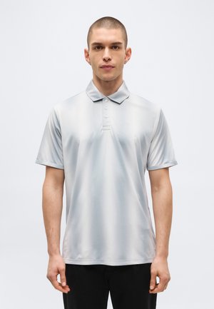 Jeune homme aux cheveux courts portant un polo gris clair à manches courtes et un pantalon noir, debout devant un fond blanc uni.