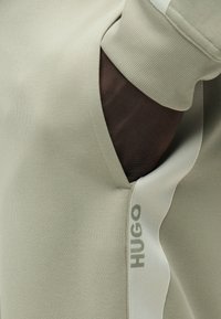 Χέρι με σκούρο χρώμα δέρματος που έχει μπει στην τσέπη ανοιχτόχρωμων μπεζ παντελονιών με ριμπ μανσέτα και λογότυπο "HUGO" στη πλευρική ραφή.