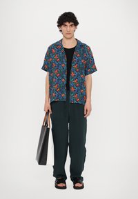 Short-sleeve shirt with a blue, green, and orange tropical print, paired with dark green trousers and black sandals, holding a black and white tote bag.