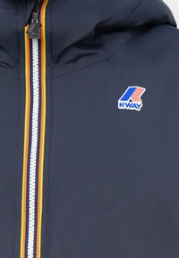 Navy K-Way jacket featuring a white zipper with red, yellow, and black stripes and a K-Way logo patch on the chest. Smooth fabric texture.