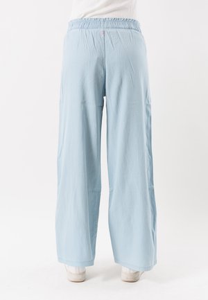 Hellblaue Hosen mit weitem Bein und elastischem Bund, getragen mit weißen Turnschuhen, von hinten auf weißem Hintergrund gezeigt.