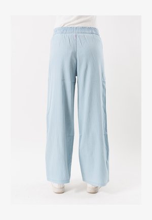 Hellblaue Hosen mit weitem Bein und elastischem Bund, getragen mit weißen Turnschuhen, von hinten auf weißem Hintergrund gezeigt.