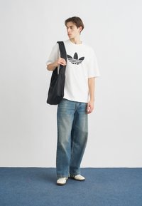 Biely oversized tričko s čiernym logom, modré široké nohavice, biele tenisky a čierna taška, stojaca na modrom koberci.