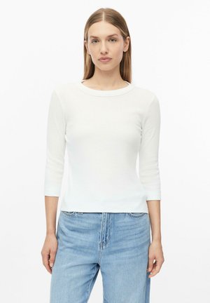 Junge Frau mit glatten hellbraunen Haaren, die ein schlichtes weißes Dreiviertelarm-Top und hellblaue Jeans trägt und vor weißem Hintergrund steht.