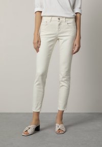 Femme portant un jean slim blanc, un haut blanc à manches retroussées et des sandales à talons ouverts beiges, se tenant sur un sol gris.