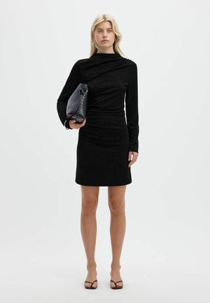 Une femme blonde porte une robe noire à manches longues et des tongs. Elle tient une pochette noire devant un fond de studio blanc uni.