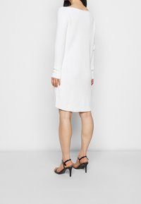 Robe blanche en maille avec manches longues, encolure arrondie et poignets côtelés, associée à des sandales à talons hauts noires à lanières.