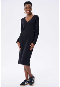 Schwarzes geripptes Kleid mit langen Ärmeln und V-Ausschnitt; figurbetonte Silhouette; kombiniert mit dunklen Sneakers mit weißen Streifen.