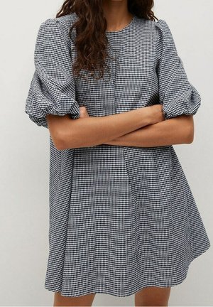 Woman with crossed arms wearing a black and white gingham puff sleeve dress with a round neckline against a plain light background.