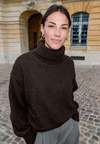 Dunkelbrauner Rollkragenpullover aus weichem, strukturiertem Material mit geripptem Kragen. Kombiniert mit hellgrauen Hosen, vor einem Kopfsteinpflaster-Hintergrund.