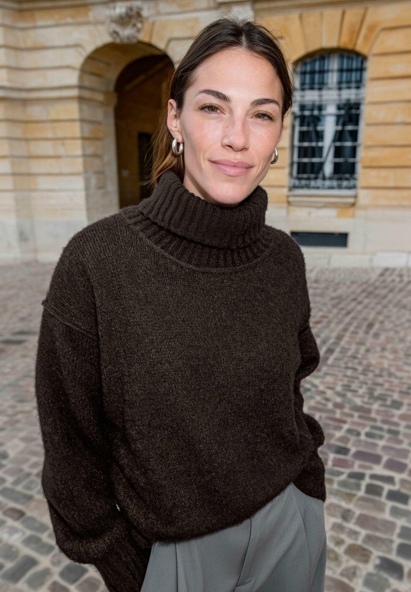 Dunkelbrauner Rollkragenpullover aus weichem, strukturiertem Material mit geripptem Kragen. Kombiniert mit hellgrauen Hosen, vor einem Kopfsteinpflaster-Hintergrund.