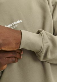 Sudadera verde oliva claro con puños acanalados. Texto "Originals Studio" impreso en blanco en el área del pecho. Textura suave, corte relajado.