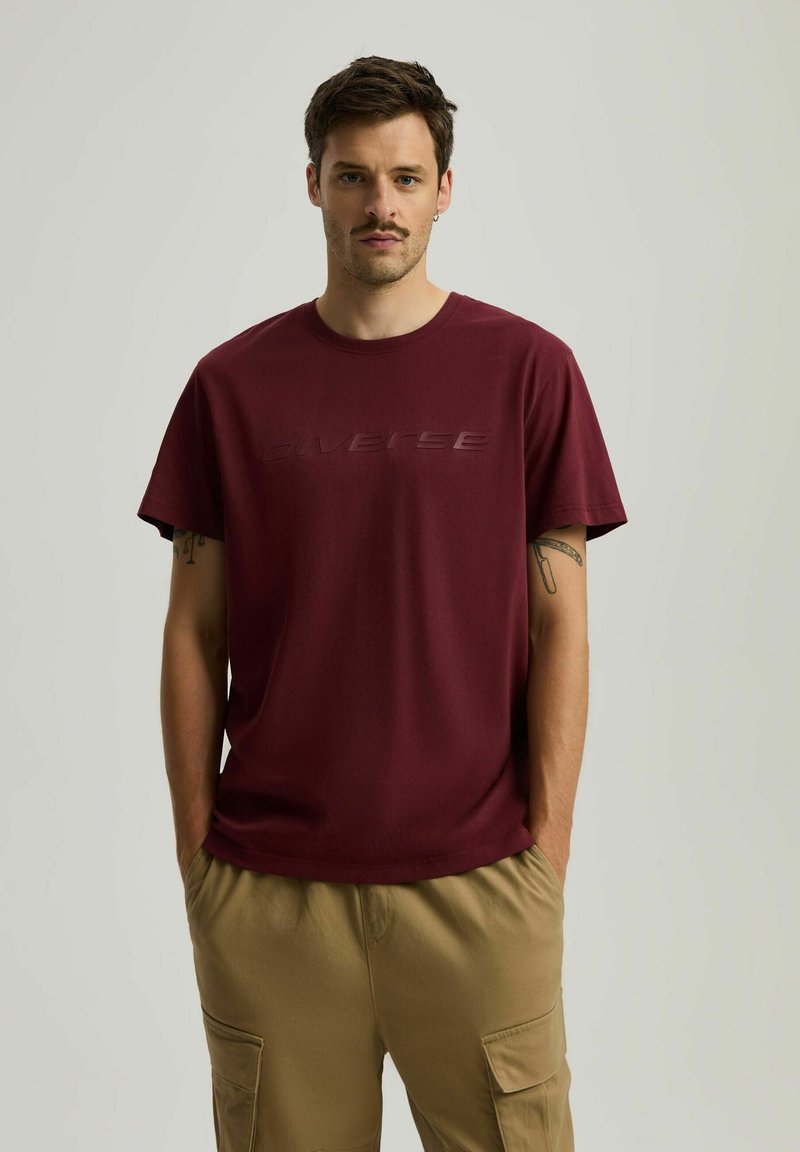 Burgundowa koszulka z okrągłym dekoltem z bawełny z wypukłym logo "Diverse" z przodu, zestawiona z beżowymi spodniami cargo. Luźny krój i krótkie rękawy.