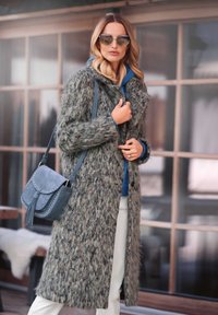 Langer, grauer, flauschiger Mantel mit strukturiertem Finish, kombiniert mit einem blauen Kapuzenpullover. Eine marineblaue Umhängetasche mit Nieten und Quastenverzierung rundet den Look ab.