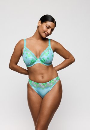 Vrouw poseert in een lichtblauwe beha van florale kant en bijpassende hoog uitgesneden slipjes tegen een effen lichte achtergrond.