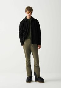 Junger Mann in einer schwarzen Fleecejacke, olivgrünen Hosen, schwarzen Winterstiefeln und großen orange getönten Sonnenbrillen, stehend vor weißem Hintergrund.