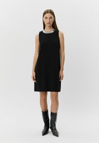 Zwart mouwloos jurkje met een ronde halslijn versierd met metalen accenten; gladde stof; gecombineerd met gestructureerde zwarte cowboylaarzen.