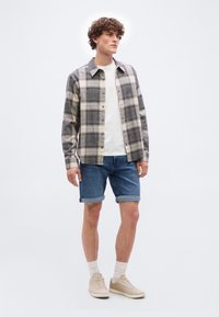 Νεαρός άνδρας με σγουρά μαλλιά που φοράει μαύρο και λευκό πουκάμισο καρό πάνω από λευκό μπλουζάκι, μπλε τζιν σορτς γυρισμένα, λευκές κάλτσες και μπεζ αθλητικά παπούτσια.