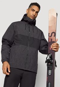 Ski jakke i svart og grå mønster med høy krage og glidelås, holdt av en person med brun hud, sammen med en ski med bindinger.