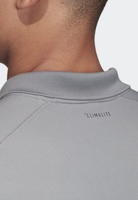 Svetle šedé sportovní tričko s texturovanou tkaninou, s plochým límečkem a textem "'CLIMALITE" vytištěným na zadní straně poblíž límečku.