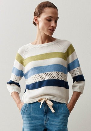 Frau trägt einen weißen Strickpullover mit grünen, blauen und marineblauen horizontalen Streifen und hochgekrempelten Ärmeln, kombiniert mit blauen Jeans und den Händen in den Taschen.