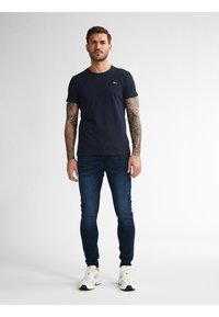 Námorně modré tričko s krátkým rukávem a kulatým výstřihem, spárované s tmavě modrými džíny a bílými teniskami. Tetování viditelné na pažích.