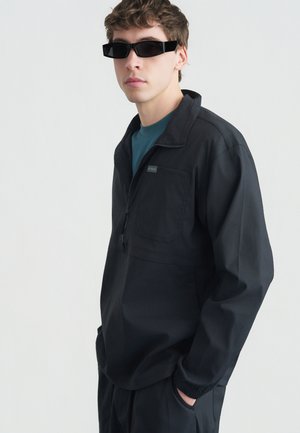 Νεαρός άνδρας που φοράει μαύρα γυαλιά ηλίου, μαύρο μπουφάν με φερμουάρ και τσέπη στο στήθος, και σκούρο παντελόνι, στέκεται μπροστά σε λευκό απλό φόντο.