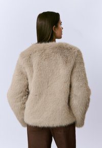 Mujer con cabello lacio castaño viste una chaqueta de piel sintética beige y pantalones marrones, de espaldas contra un fondo gris claro liso.