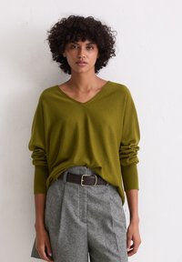 Oliwkowy zielony sweter w serek wykonany z miękkiego materiału, zestawiony z szaro-skalowym, wysokim w pasie spodniami w kratę oraz ciemnobrązowym paskiem z złotą klamrą.