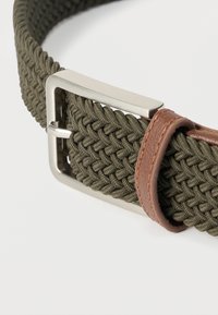 Πλεκτή ζώνη από υφασμάτινο ελιάς πράσινου χρώματος με ασημένια ορθογώνια αγκράφα και καφέ δερμάτινο λουράκι, σε λευκό φόντο.