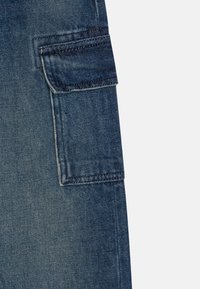 Sekce džínových cargo kalhot s velkou kapsou na klopě, tmavě modrá barva, hrubá textura a viditelné švy podél okrajů.