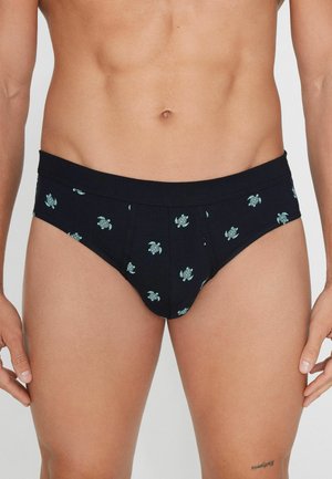 Torso masculino con calzoncillos negros con pequeños patrones de tortugas en azul claro sobre un fondo claro liso.