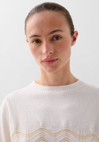 Cremefarvet strikket sweater med ribbet halsudskæring og bølget mønsterdetaljer i lysere nuancer, med en blød tekstur og afslappet pasform.