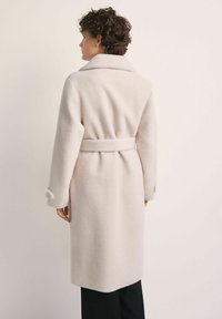 Osoba z krótkimi kręconymi włosami nosi jasnobeżowy wełniany płaszcz z paskiem, stojąc tyłem na tle w jednolitym jasnym kolorze.