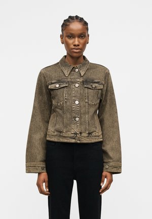 Žena oblečená v vyblednutej olivovo zelenej rifľovej bunde s prednými vreckami a striebornými gombíkmi, v kombinácii s čiernymi nohavicami, stojaca pred jednoduchým pozadím.