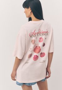 T-shirt oversize rose clair avec un motif graphique de coquillages multicolores et du texte au dos. Manches courtes, tissu doux.