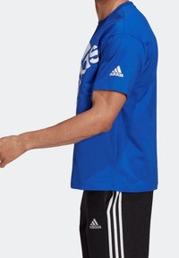 Modrá bavlněná tričko s bílým logem Adidas na hrudi, krátké rukávy a kulatý výstřih. Nosené s černými tepláky se třemi pruhy.