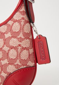 Coin de sac à main rouge et beige à motifs avec bordure en cuir rouge, chaîne argentée et étiquette en cuir rouge embossée du nom de la marque « Coach ».