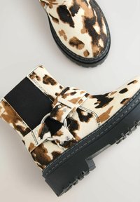 Botine cu imprimeu de vacă, fabricate dintr-un material texturat, cu o parte laterală elastică neagră, detalii din metal argintiu și o talpă groasă din cauciuc.