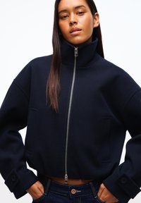 Pitkähiuksinen nainen, jolla on ylhäältä vetoketjullinen tummansininen lyhyt takki ja tummansiniset farkut, kädet taskuissa, neutraali ilme.