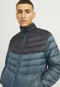Giacca a vento con colletto alto, caratterizzata da pannelli trapuntati orizzontali in blu scuro e teal. Chiusura con zip e logo discreto sulla manica.