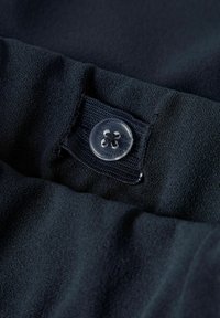 Mørkeblåt stof med en glat overflade. Har en klar knap og et elastisk bånd til fastgørelse, syet ind i tøjmaterialet.