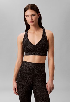 Femme portant un brassard de sport noir à motifs et un legging taille haute, debout devant un fond gris uni, visage détendu regardant vers l'avant.