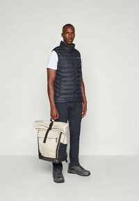 Melna pūkainā veste virs baltas kreklas, kopā ar tumšām biksēm un melniem apaviem. Beigēta mugursoma ar melniem akcentiem, turēta vienā rokā.