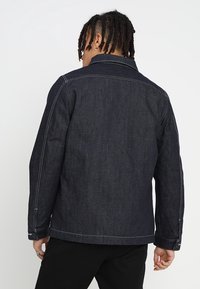 Tmavá džínová bunda s přímým střihem, viditelnými švy a klasickým límcem. Zádní část má jednoduchý, minimalistický design.