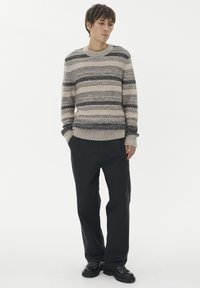 Ung man med händerna i fickorna iförd en beige och svart randig stickad tröja, svarta byxor och svarta snörkängor, stående mot en vit bakgrund.