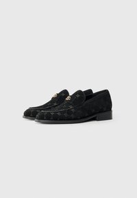 Mocassins en daim noir avec un motif, des éléments décoratifs en or et un petit talon carré. Texture lisse avec des détails de couture.