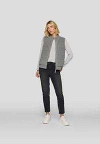 Gray quilted vest with textured front and two pockets, worn over a light gray long-sleeve shirt, paired with black jeans and white sneakers.