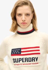 Donna con capelli castano chiaro che indossa un maglione a collo alto crema con la bandiera americana e il testo "Superdry Authentic Branded Goods" sul davanti.
